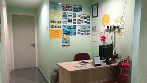 A reception desk with a computer, a fire extinguisher, and a chair. There are posters on the wall and a door with a number. - Best Seven Motel (Langkawi)