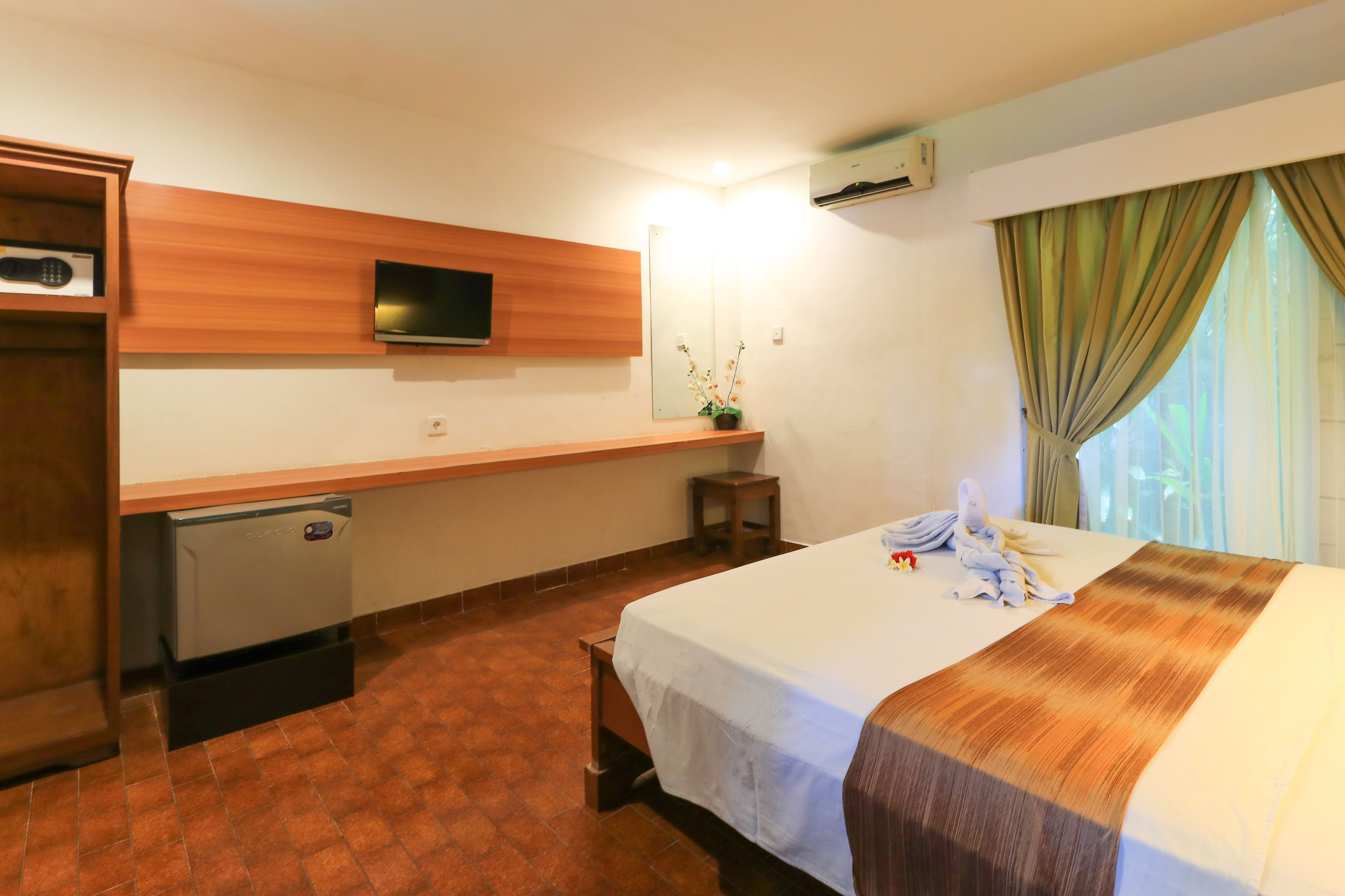 A hotel room with a bed, a television mounted on the wall, a small refrigerator, and a window with curtains.