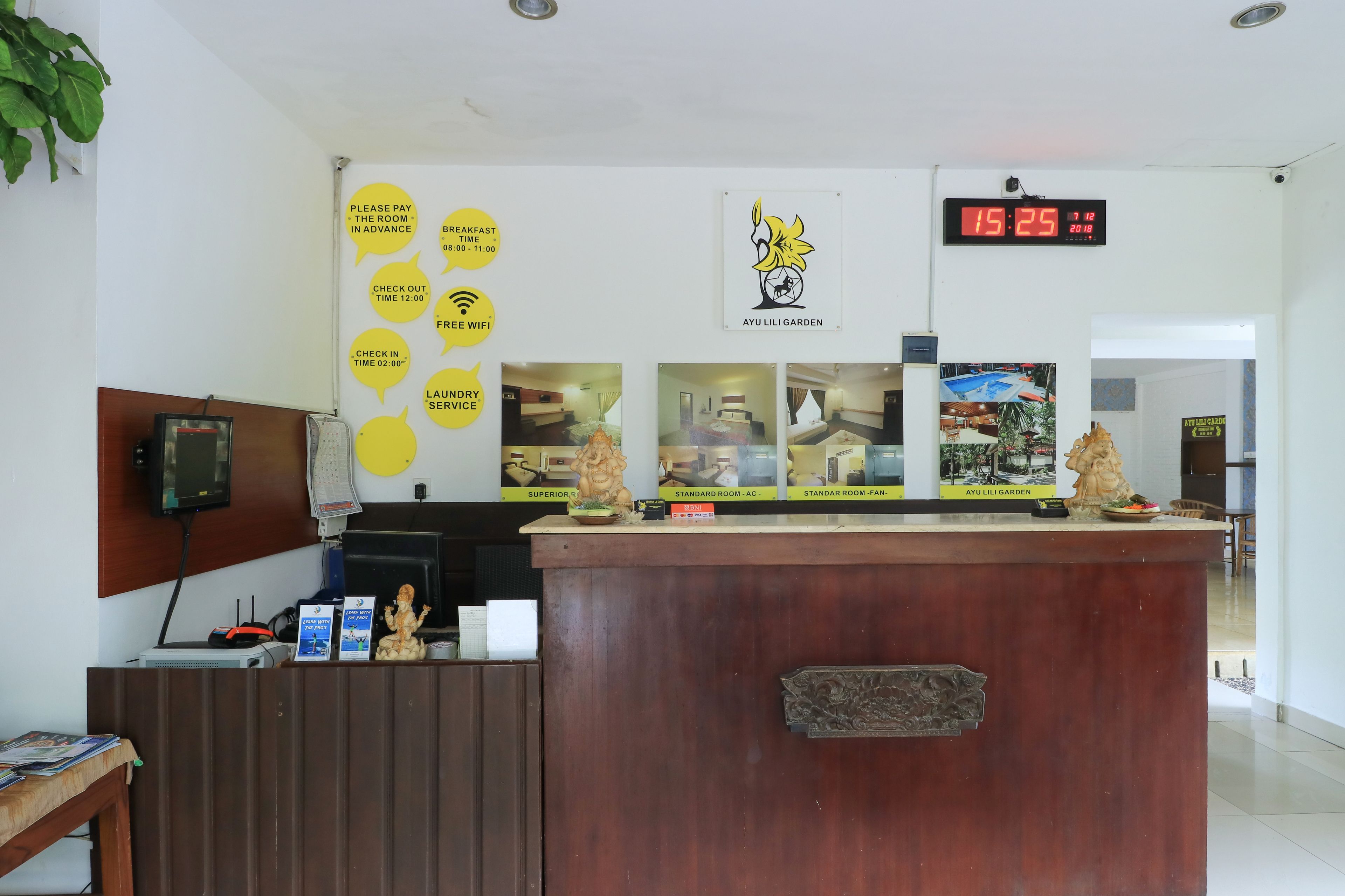 Un comptoir d’accueil d’hôtel avec une horloge numérique affichant 15h25, une enseigne indiquant le « Jardin Ayu Lili », ainsi que divers panneaux d’information sur les heures d’enregistrement et les services proposés.