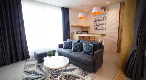 A modern living room with a grey sofa, a round coffee table, and a patterned rug. There are two pendant lights above the sofa and a kitchen area in the background.