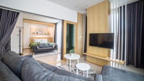 Un salon moderne avec un canapé d’angle gris, un téléviseur à écran plat fixé à un mur en bois et une porte-fenêtre coulissante donnant sur une chambre.