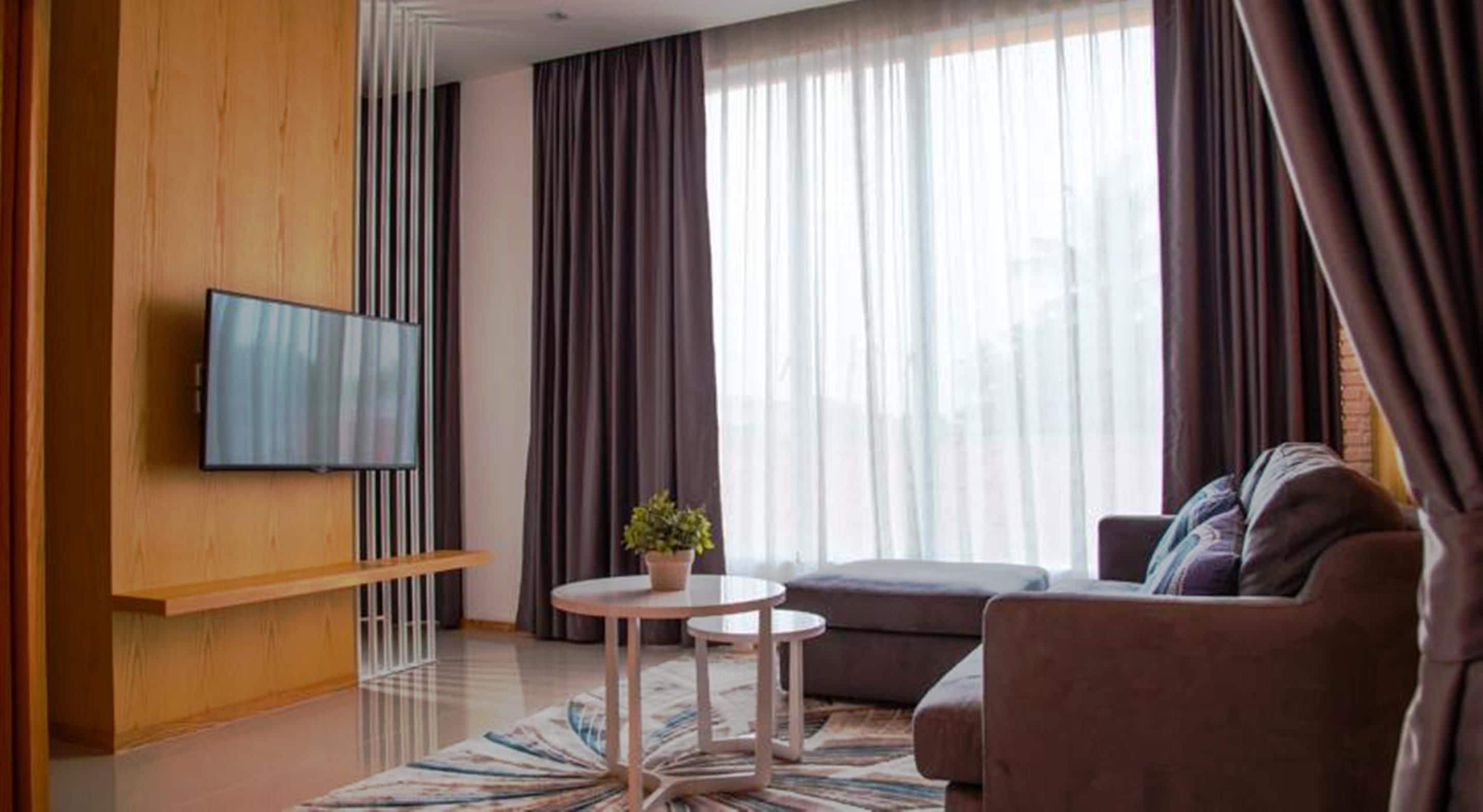 A modern living room with a flat-screen TV mounted on a wooden panel, a brown sofa, a small round table with a plant, and large windows with dark curtains.