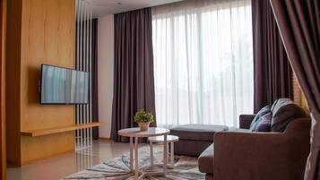 A modern living room with a flat-screen TV mounted on a wooden panel, a brown sofa, a small round table with a plant, and large windows with dark curtains.