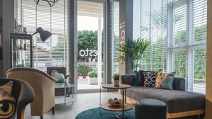 Un salon moderne avec un canapé, un fauteuil et une table basse ronde, de grandes fenêtres équipées de stores et une vue sur l’extérieur.