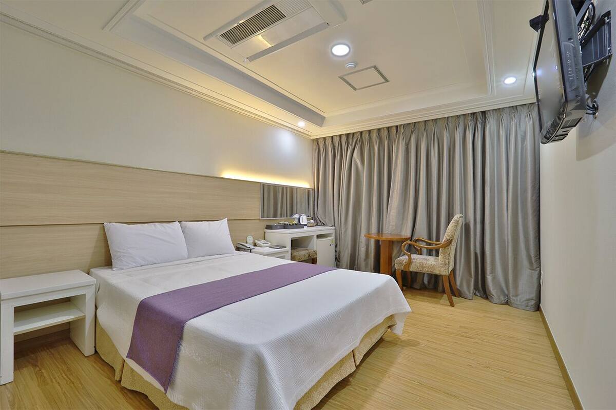 A hotel room with a large bed, bedside tables, a desk, a chair, and a TV mounted on the wall.