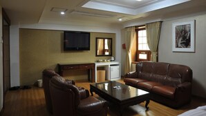 A hotel room with a brown leather sofa, a glass coffee table, a desk with a computer, a flat-screen TV, and a bed.