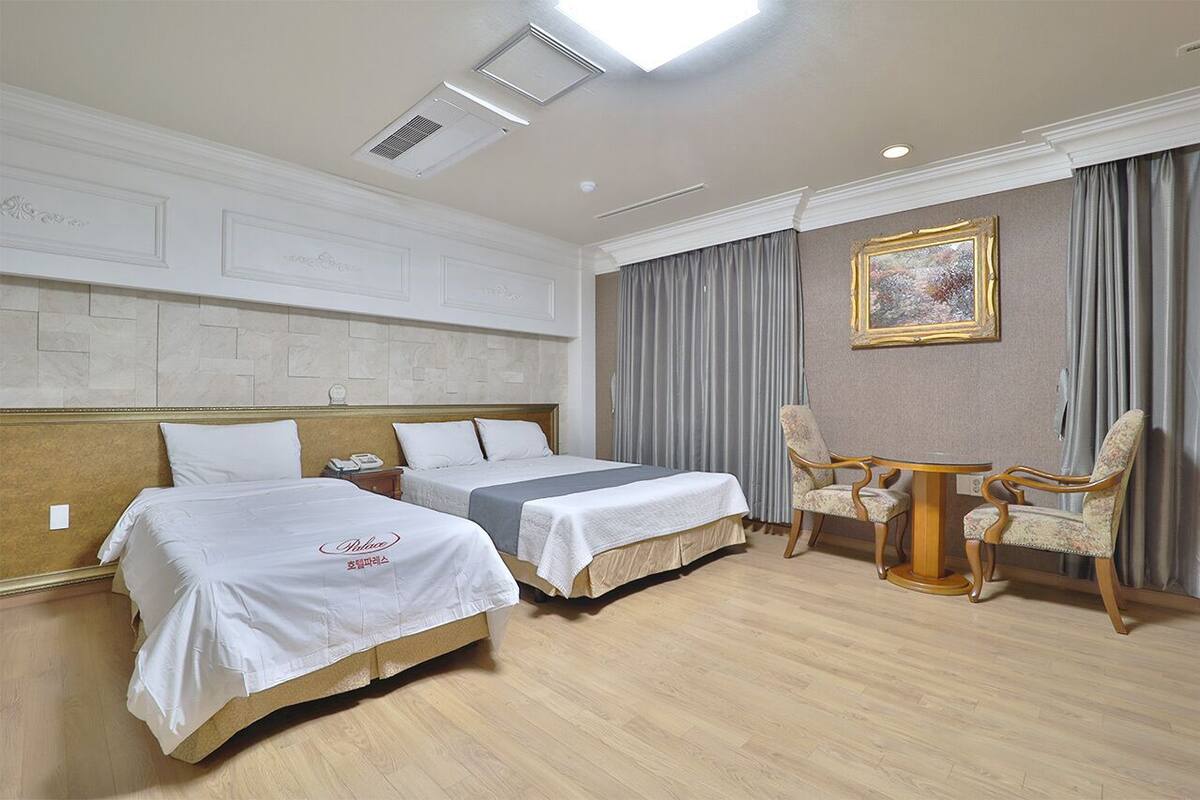 A hotel room with two beds, a small table with chairs, a painting on the wall, and a ceiling light.