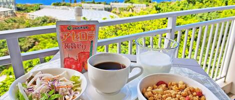 A table with a salad, coffee, milk, and a snack bowl on a balcony with a view of the sea.