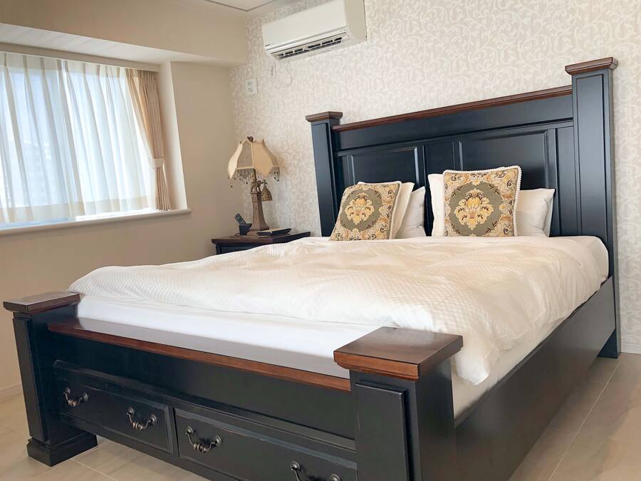 A neatly made bed with a dark wooden headboard, a bedside table with a lamp, and a wall-mounted air conditioner.