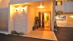 Una entrada moderna de edificio con fachada de piedra, una puerta y un pequeño jardín con plantas.