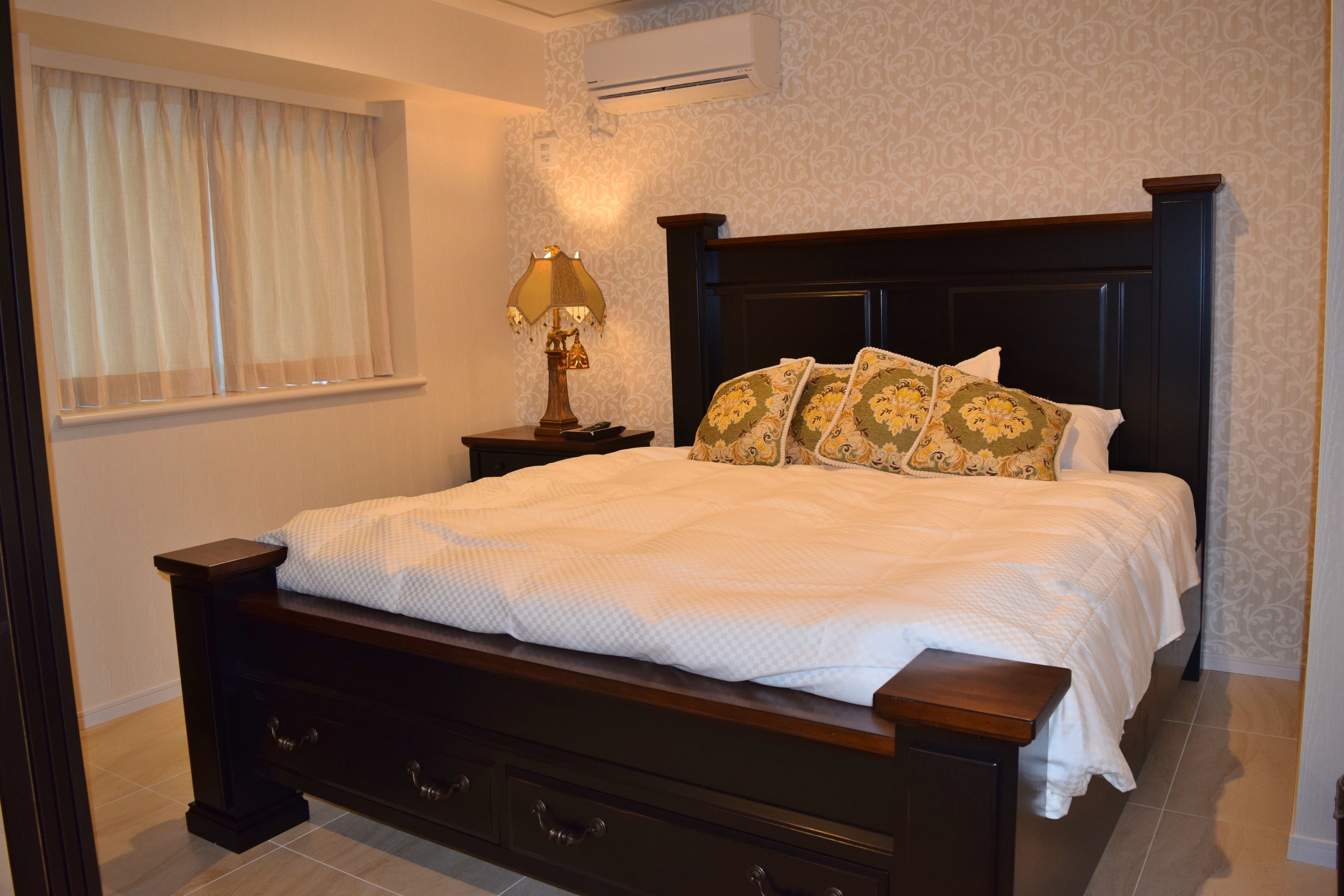 A neatly made bed with a white bedspread, a dark wooden headboard, and decorative pillows. There is a bedside table with a lamp and a wall-mounted air conditioner above the bed.