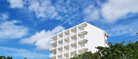 Un edificio blanco de varios pisos con balcones, rodeado de árboles y bajo un cielo parcialmente nublado.