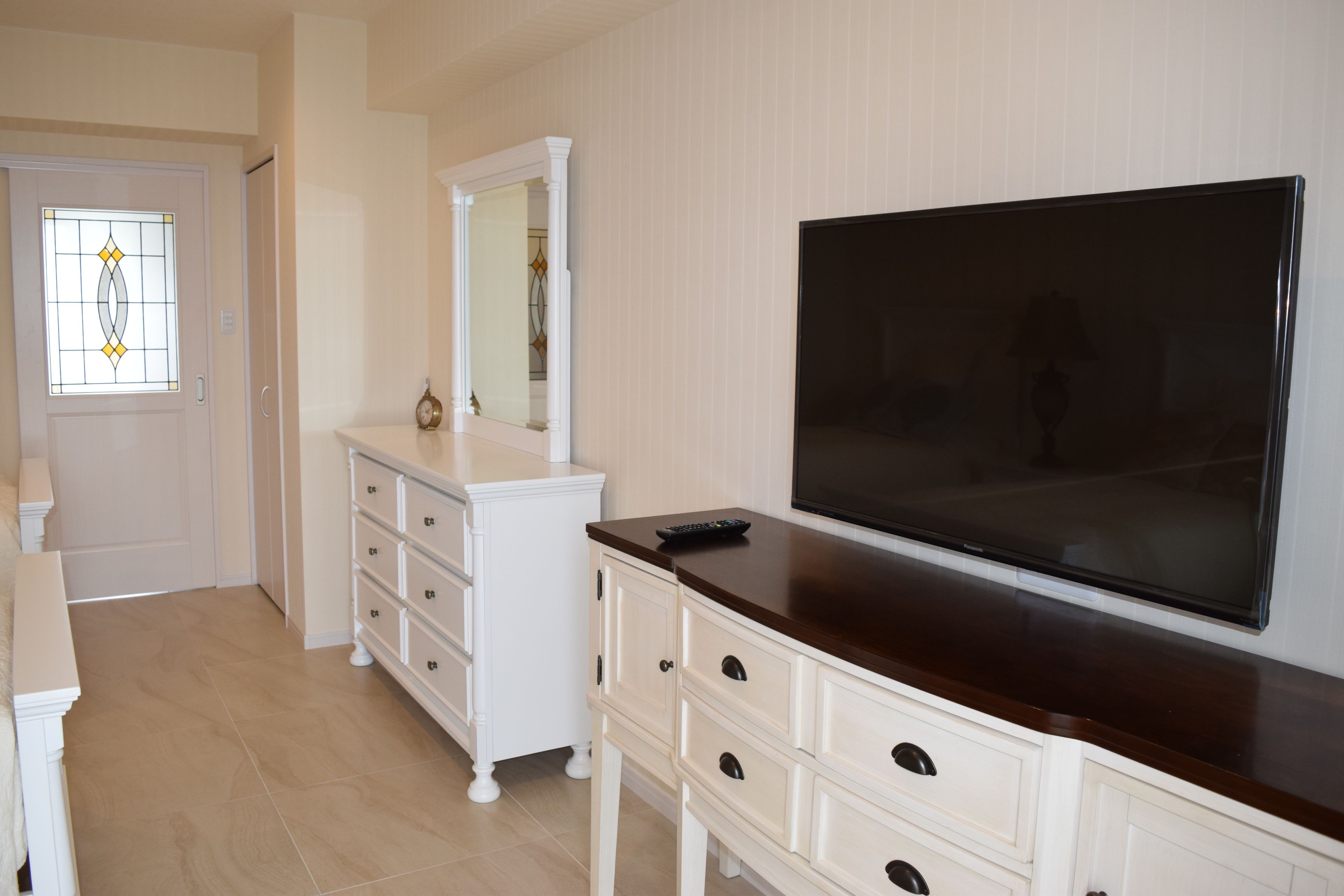 A bedroom with a large flat-screen TV mounted on the wall, a white dresser with drawers, and a door with a stained glass window.