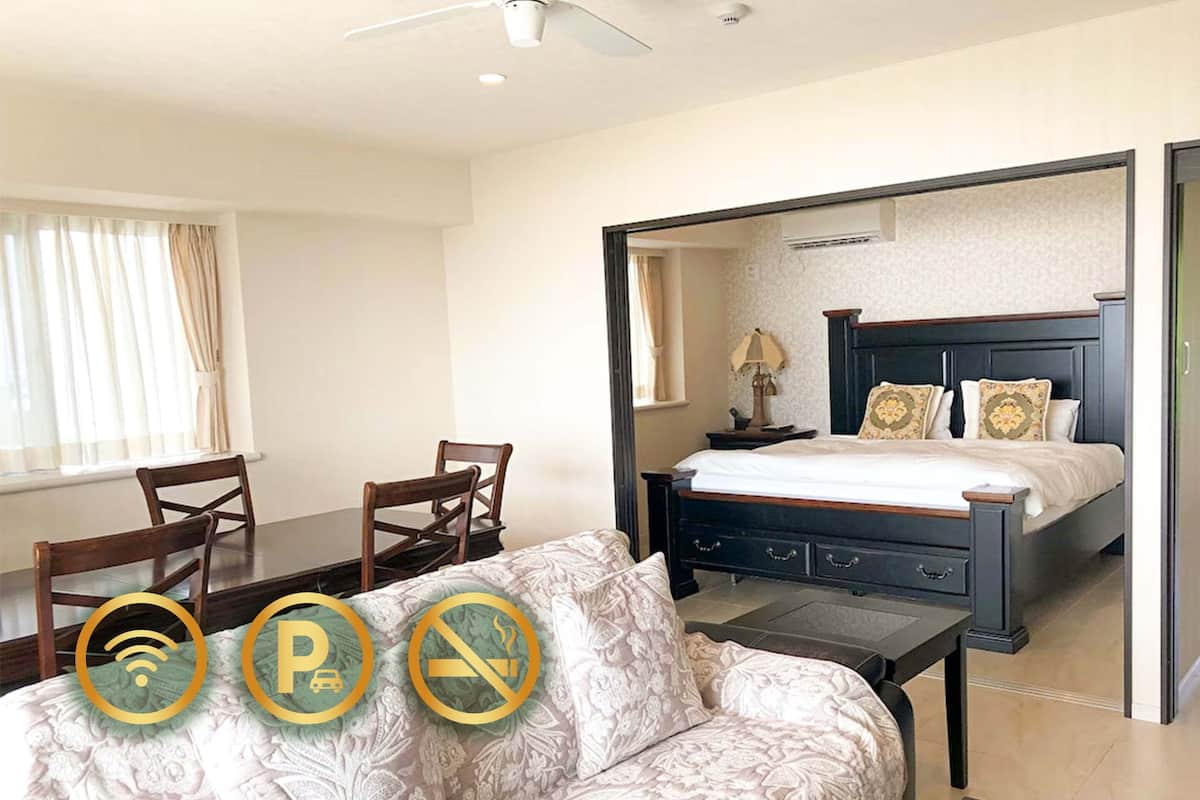 A hotel room with a bed, a sofa, a desk, and a chair. There is a window with curtains, a ceiling fan, and an air conditioning unit.