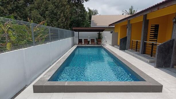 Ein rechteckiger Pool mit einer gefliesten Liegefläche, umgeben von einer weißen Mauer und einem Metallzaun.