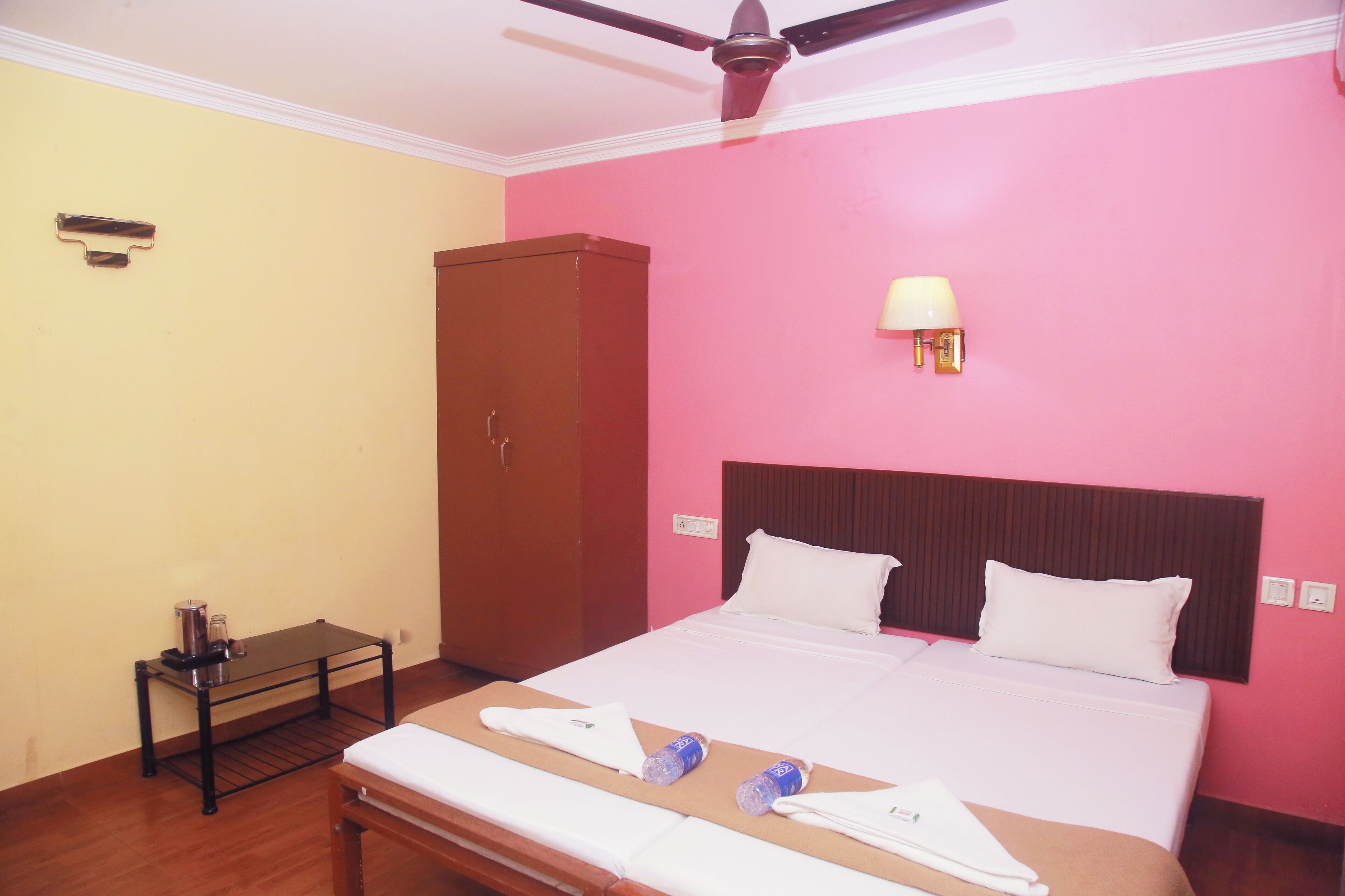 A hotel room with a bed, a wooden wardrobe, a small table with a lamp, and a ceiling fan.