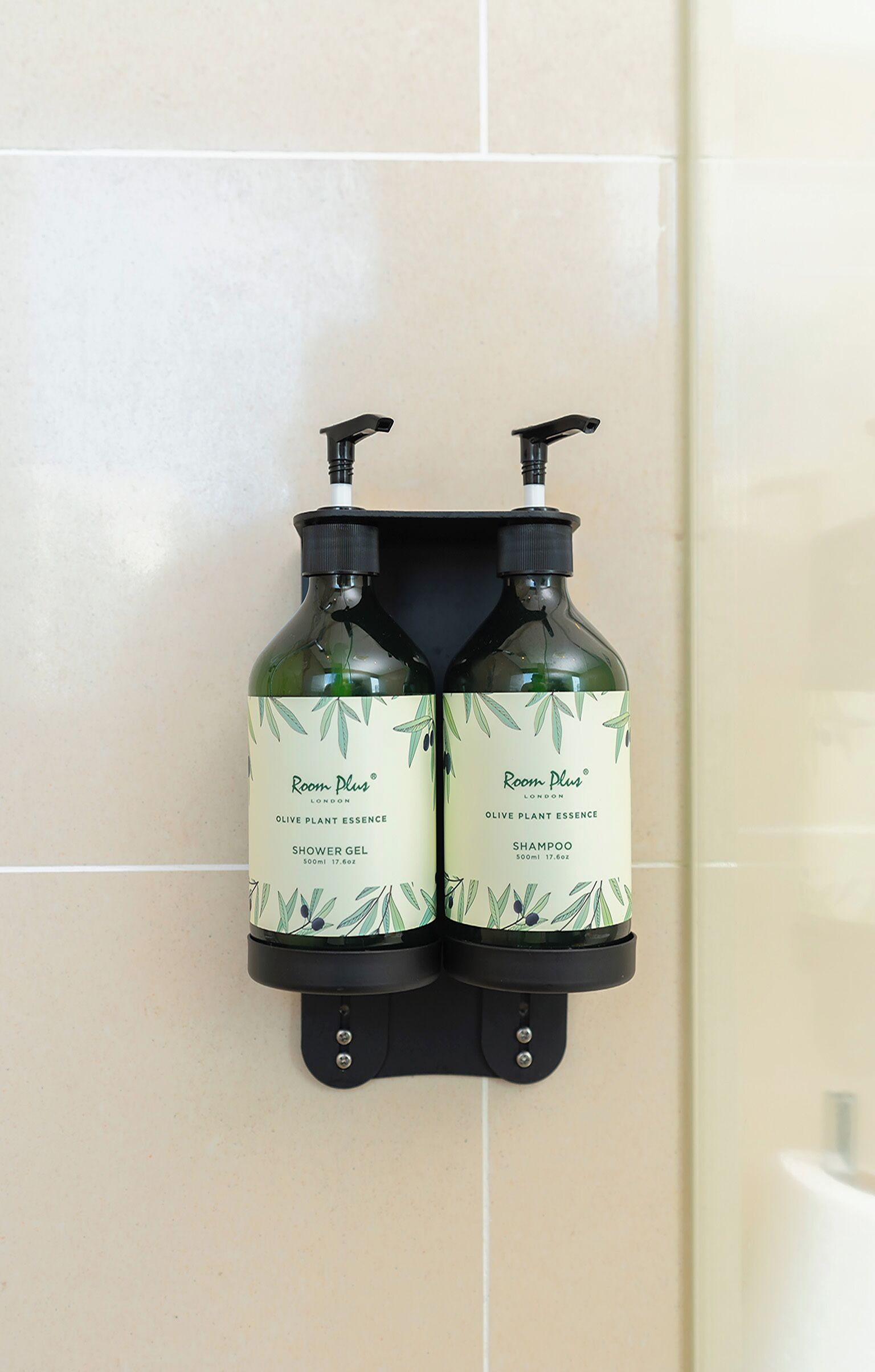 Two bottles of Room Plus Olive Plant Essence shower gel and shampoo mounted on a wall.