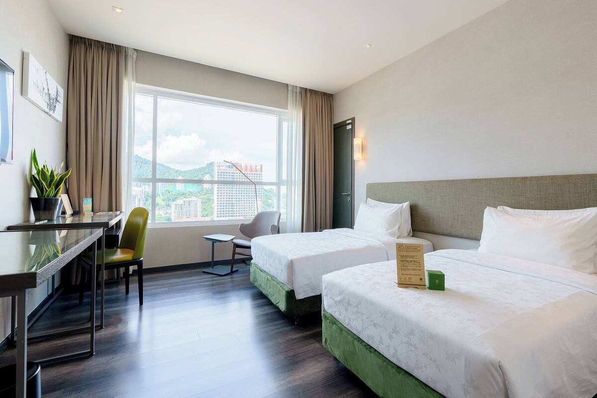 A hotel room with two beds, a desk, a chair, a potted plant, and a view of buildings through the window.