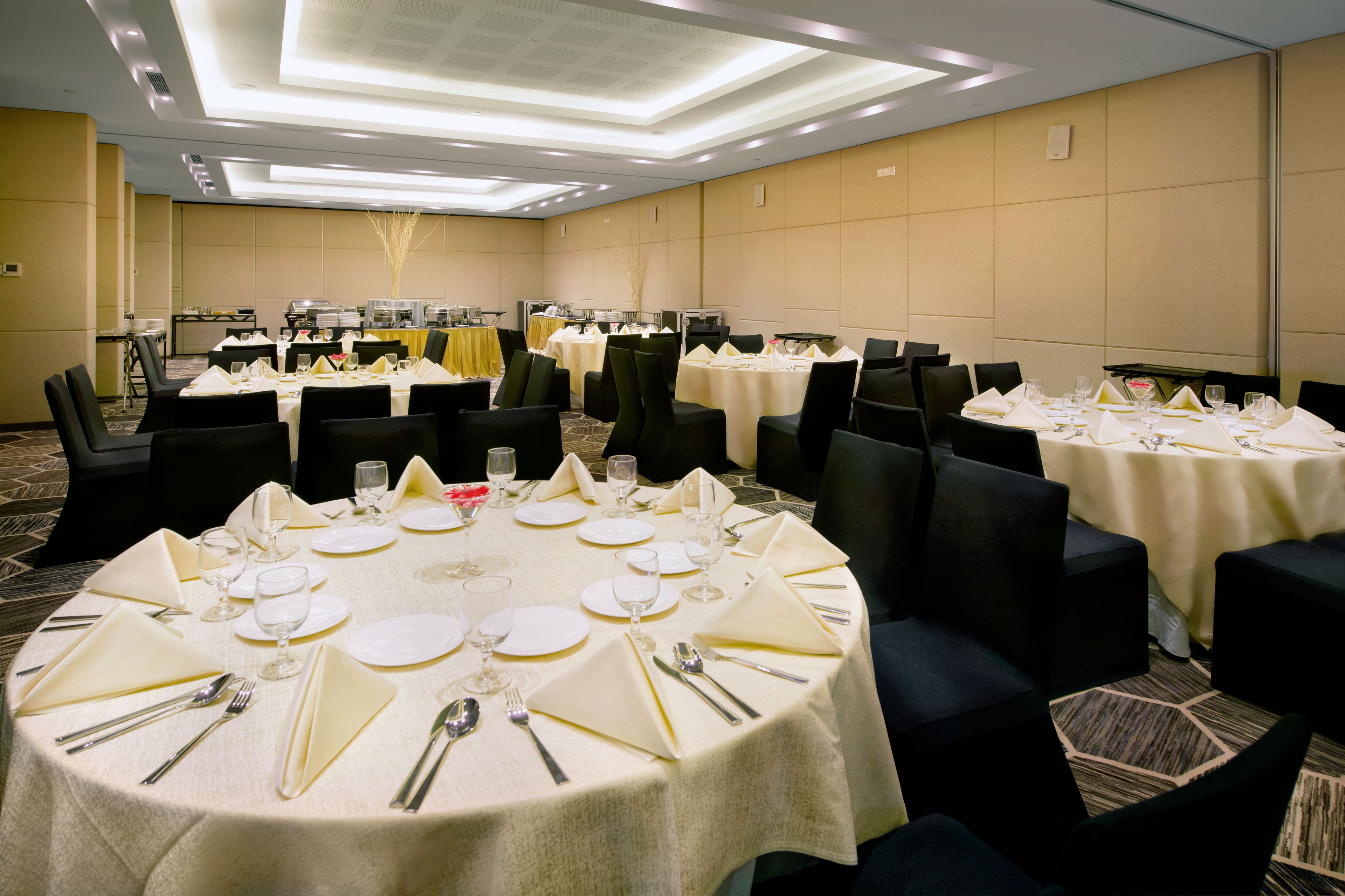 A banquet hall with round tables set for dining, equipped with chairs, and a buffet area in the background.