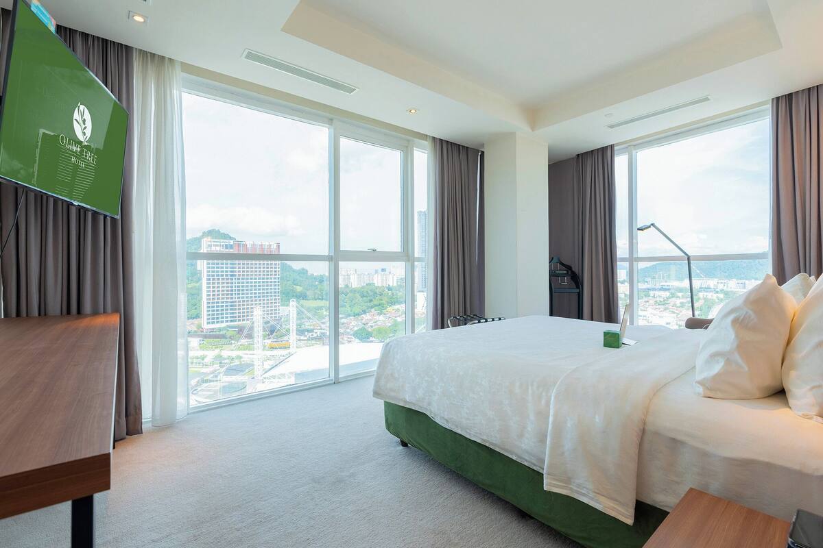 A hotel room with a large bed, a desk, and a TV. There are large windows with a city view.