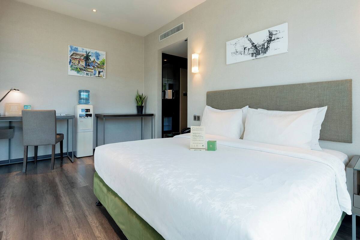 A hotel room with a bed, a desk, a chair, a water dispenser, and two artworks on the walls.