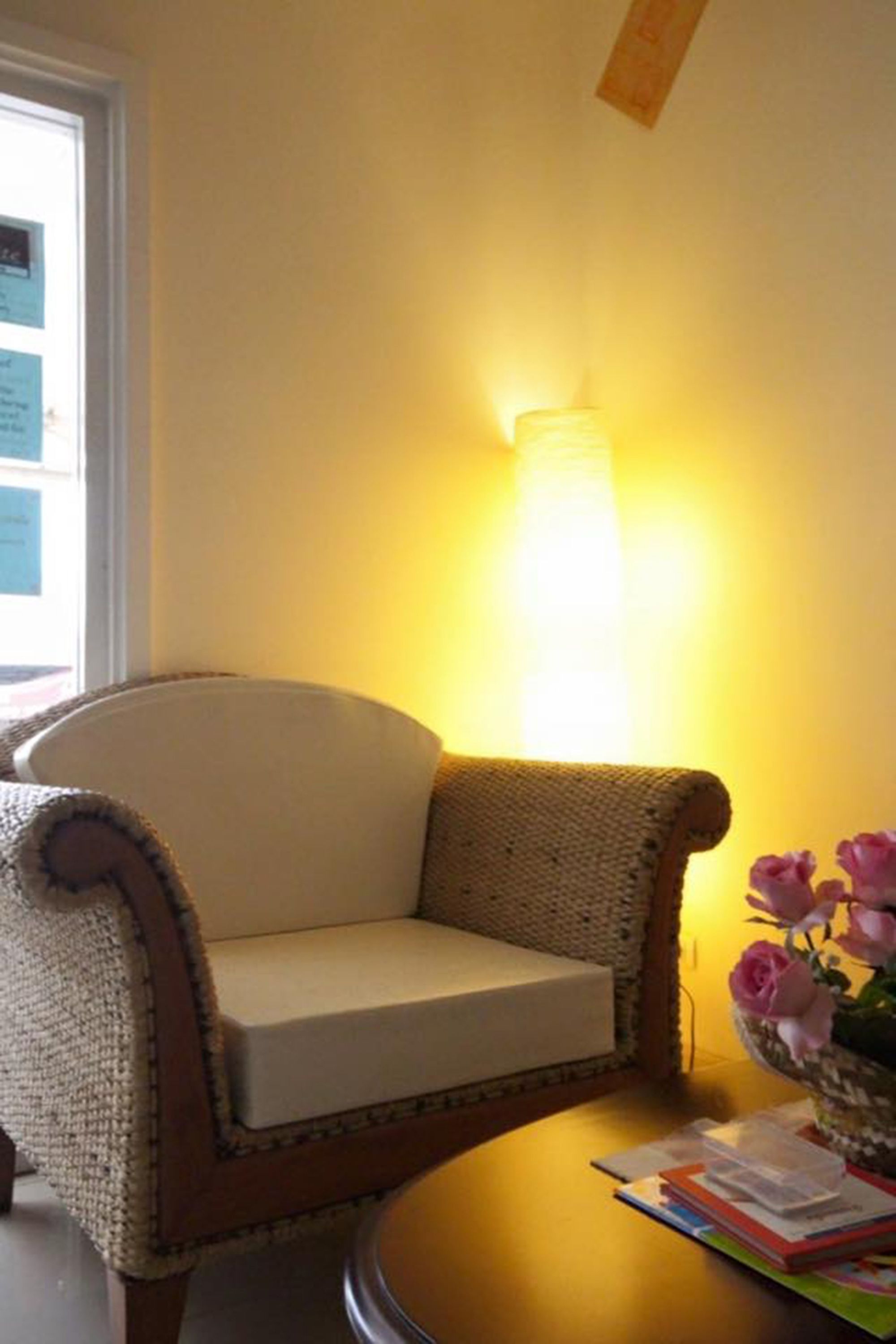 A cozy room with a wicker armchair, a small round table with a vase of flowers, and a wall-mounted lamp.