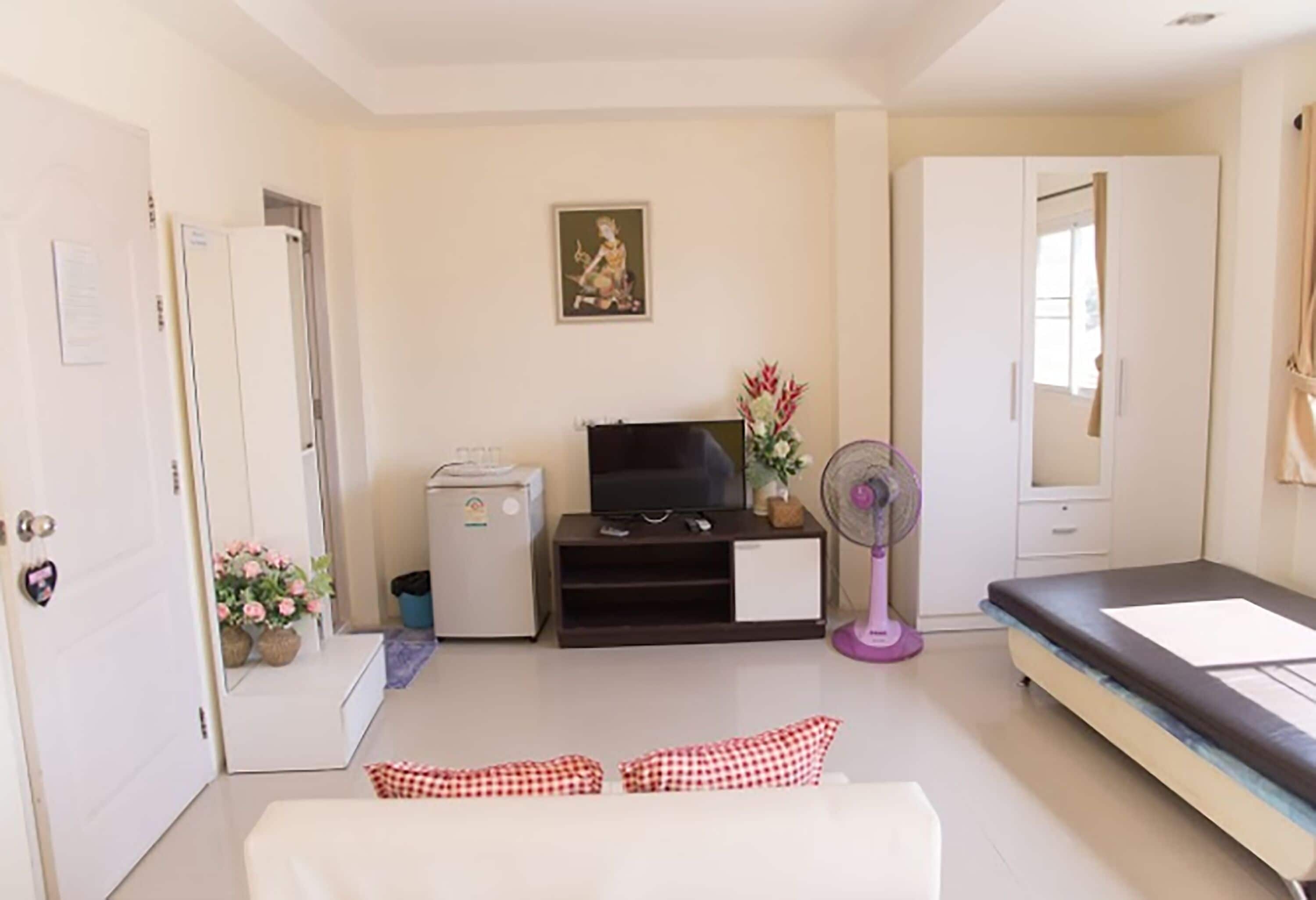 A compact living space with a sofa, a television, a small refrigerator, a fan, and a bed.