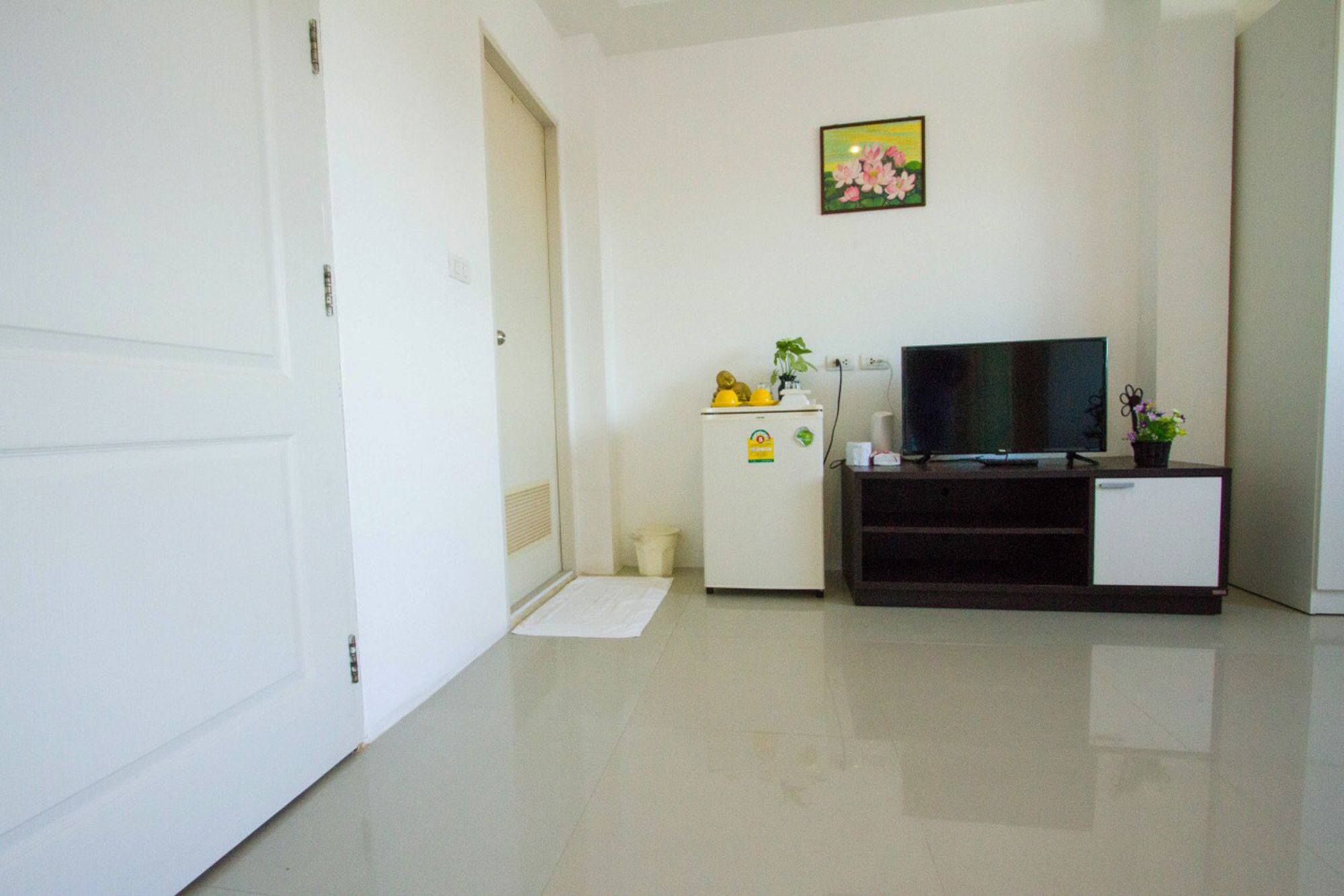 A small, clean room with a white door, a white refrigerator, a television on a stand, and a small table with a plant.