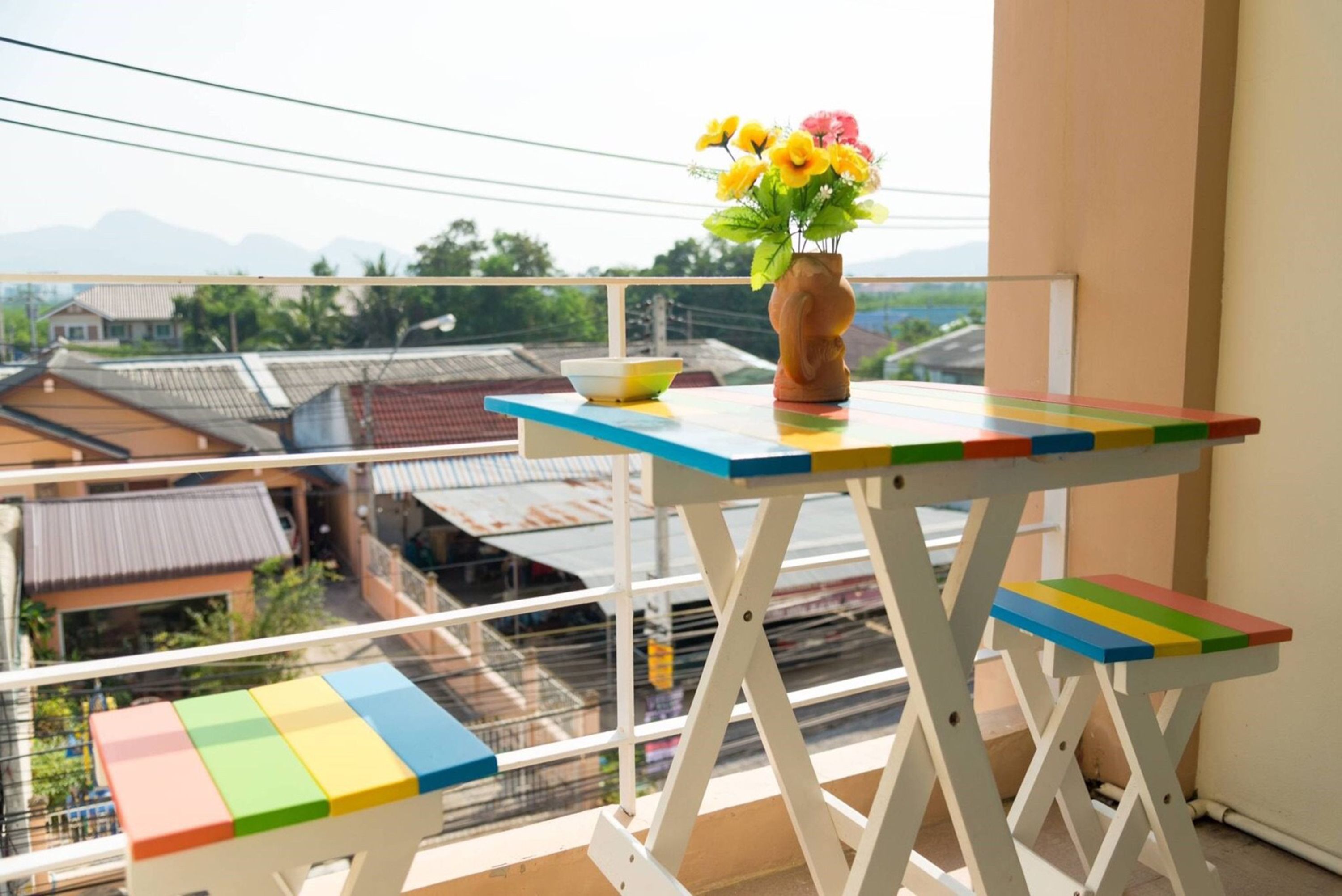 Ein Balkon mit kleinem Tisch und Bank, einer Blumenvase und Blick auf Gebäude und Berge.