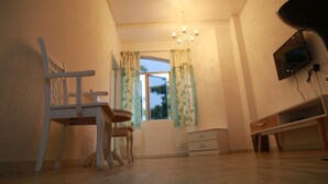A room with a wooden floor, a white chair, a small table, a TV mounted on the wall, and a window with curtains. - Thanks Hotel (Nibong Tebal)