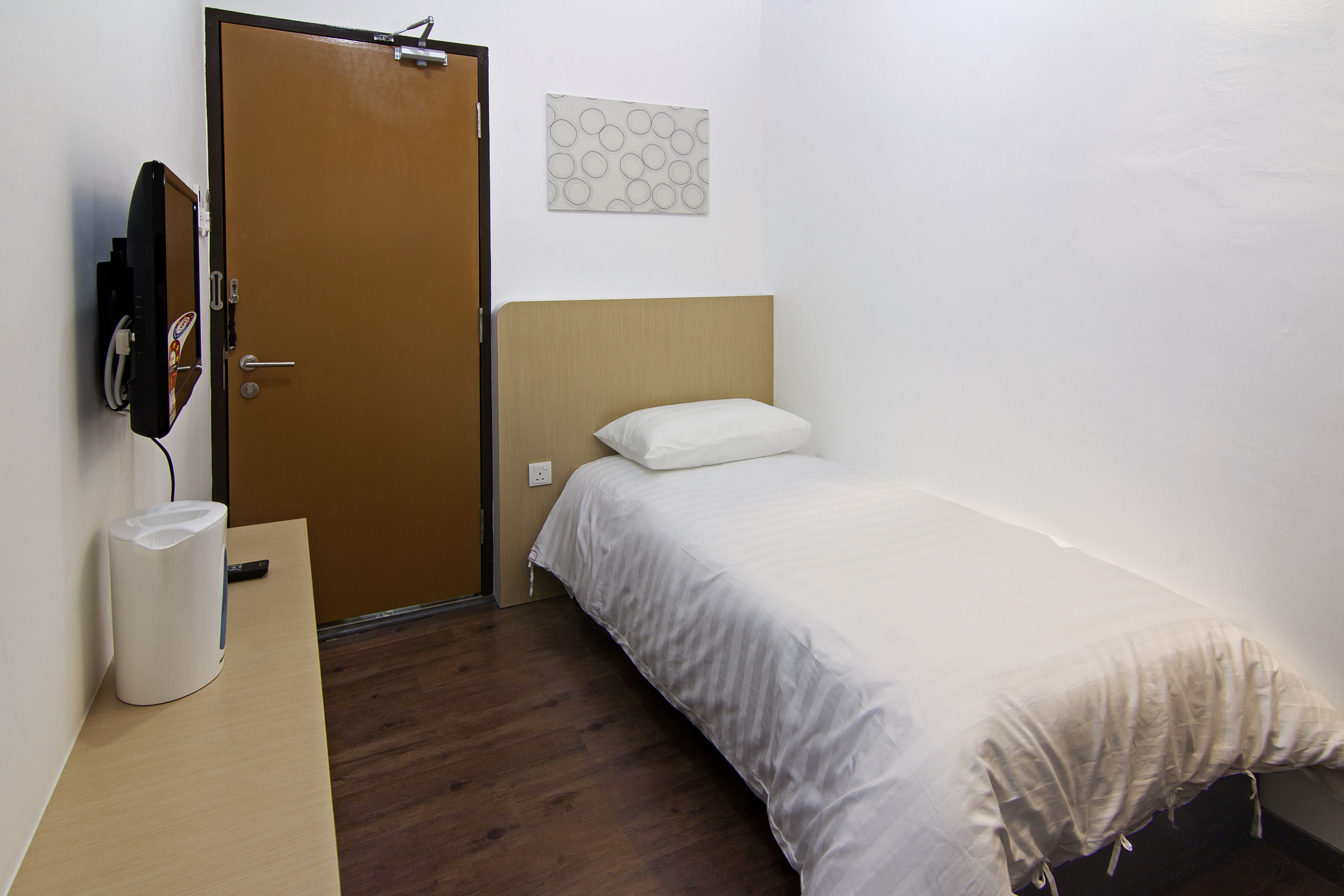 A small, neatly arranged hotel room with a single bed, a bedside table, a wall-mounted TV, and a door.