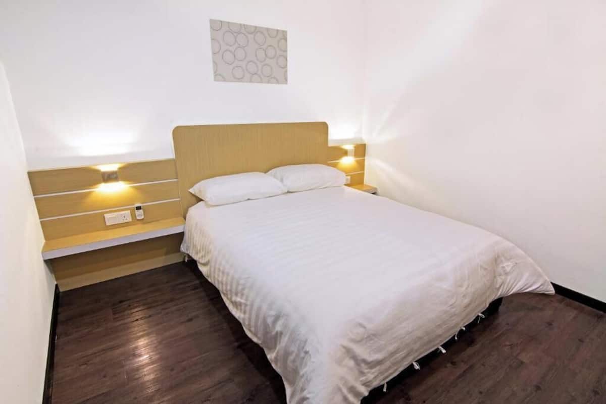 A single bed with a wooden headboard, two bedside lamps, a wall-mounted power outlet, and a patterned wall decoration.