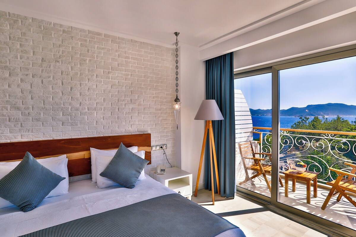 A hotel room with a white brick wall, a wooden headboard, a bed with white and blue bedding, a bedside table with a lamp, and a balcony with a view of the sea and mountains.
