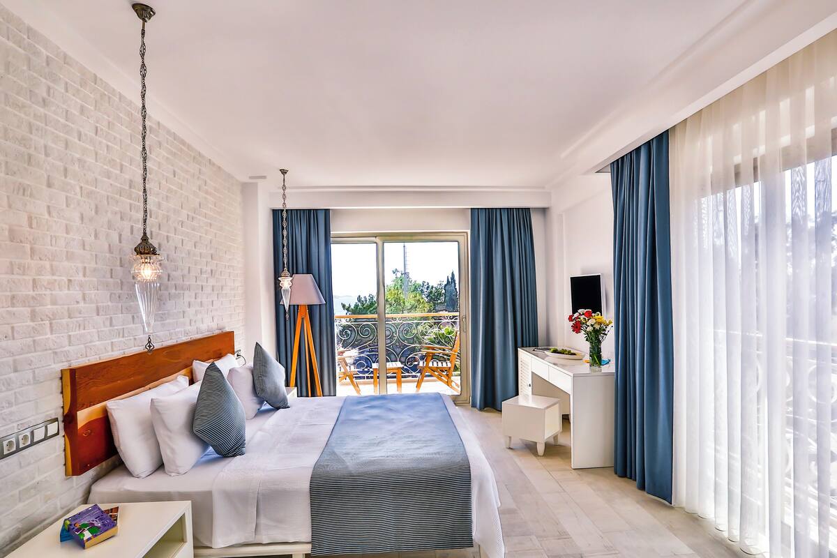 A modern hotel room with a large bed, a desk, and a balcony with blue curtains.