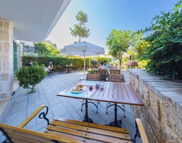 Un patio al aire libre con mesas y bancos de madera, rodeado de follaje y muros de piedra.