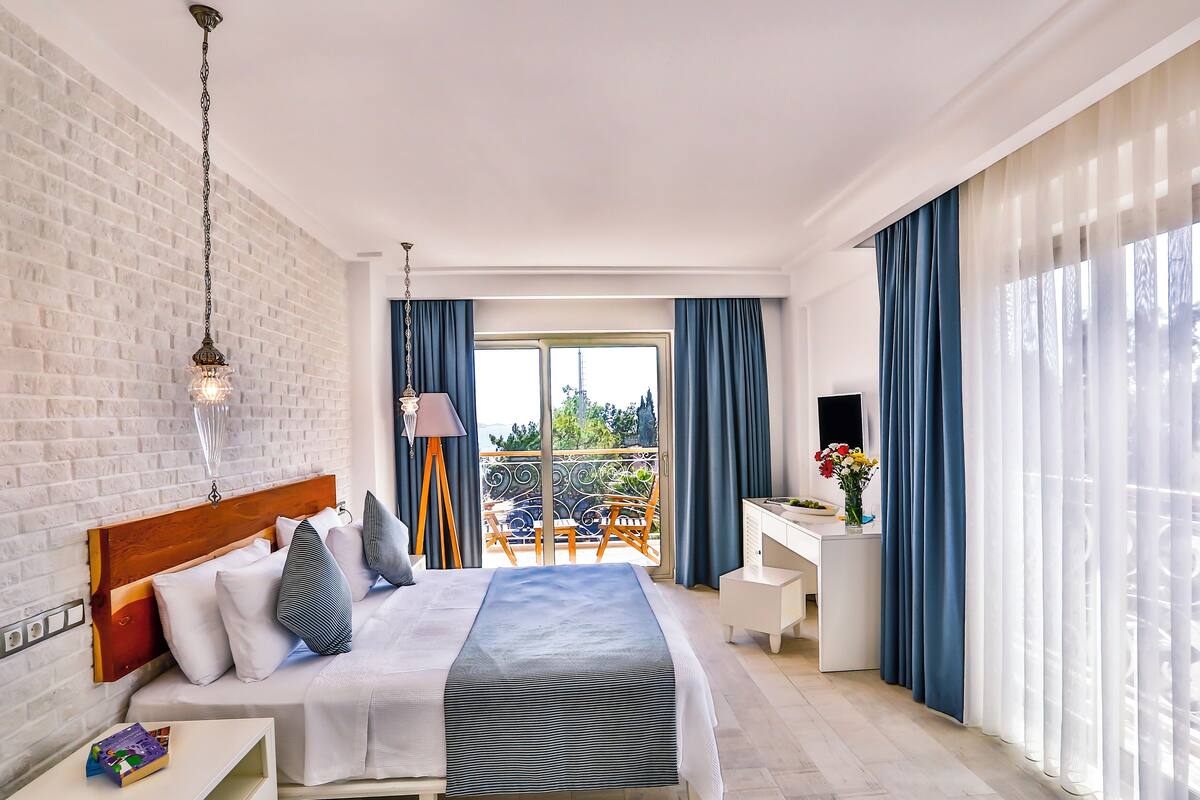 A modern hotel room with a large bed, a desk, and a balcony with blue curtains.