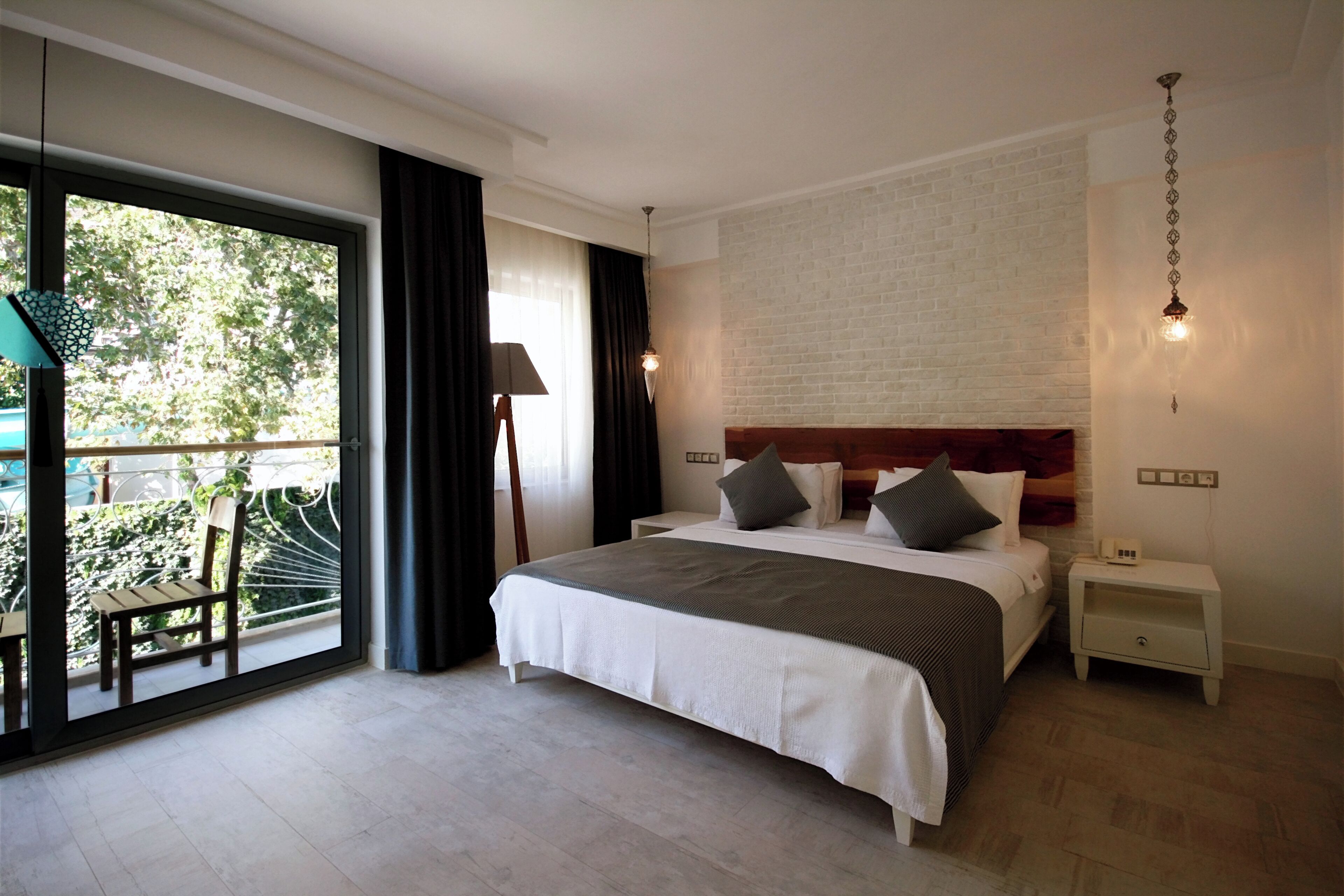 A bedroom with a large bed, a nightstand, a lamp, a view of greenery through a sliding door, and a brick wall.