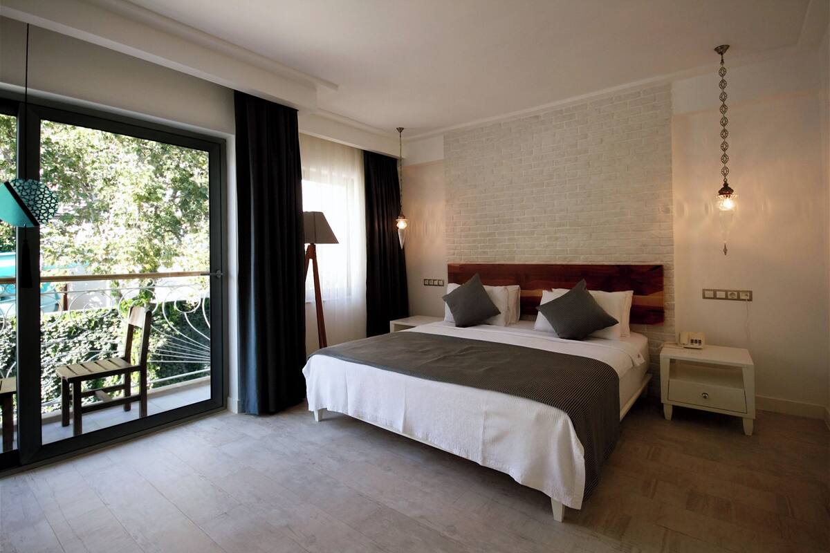 A bedroom with a large bed, a nightstand, a lamp, a view of greenery through a sliding door, and a brick wall.
