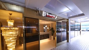 Exterior del hotel Den Basta, con un diseño moderno, que presenta una entrada de cristal, un candelabro y un estacionamiento visible al fondo.