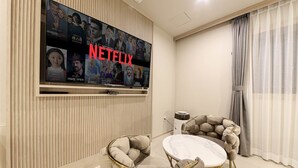 Una sala de estar moderna con un televisor de pantalla plana grande montado en la pared mostrando contenido de Netflix. Hay una mesa de mármol redonda con dos sillas y un cojín decorativo en el suelo.