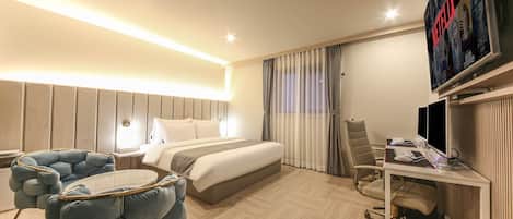 A hotel room with a bed, a desk with a computer, a TV, and a seating area with a chair and a table.