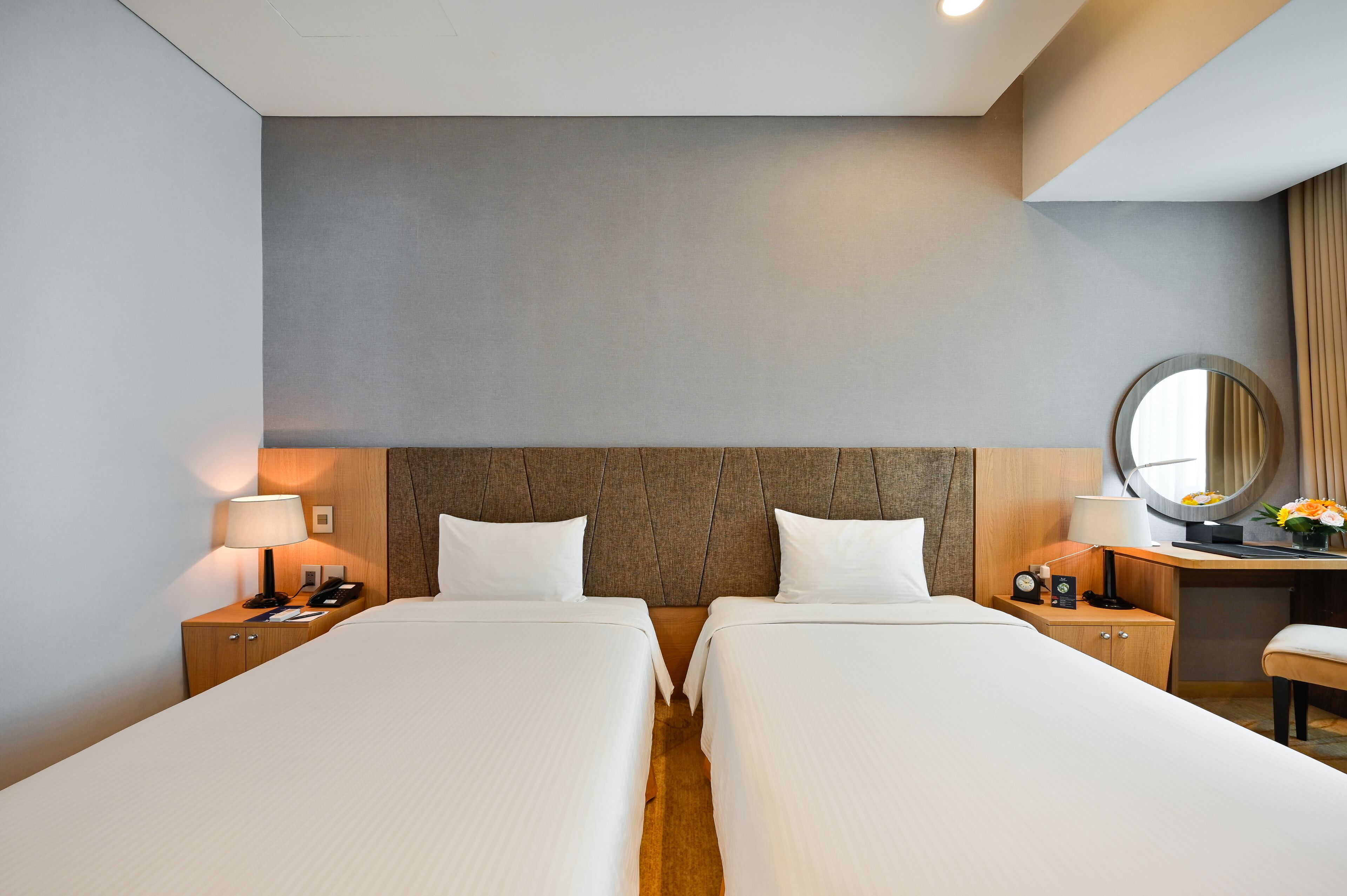 Una habitación de hotel con dos camas, un cabecero de madera, un escritorio con una silla, un espejo redondo y una mesita de noche con una lámpara.