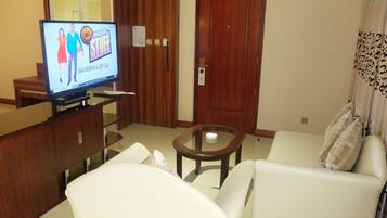 Habitación de hotel con televisión mostrando un programa deportivo, una puerta de madera, un sofá y una mesa de centro de vidrio.