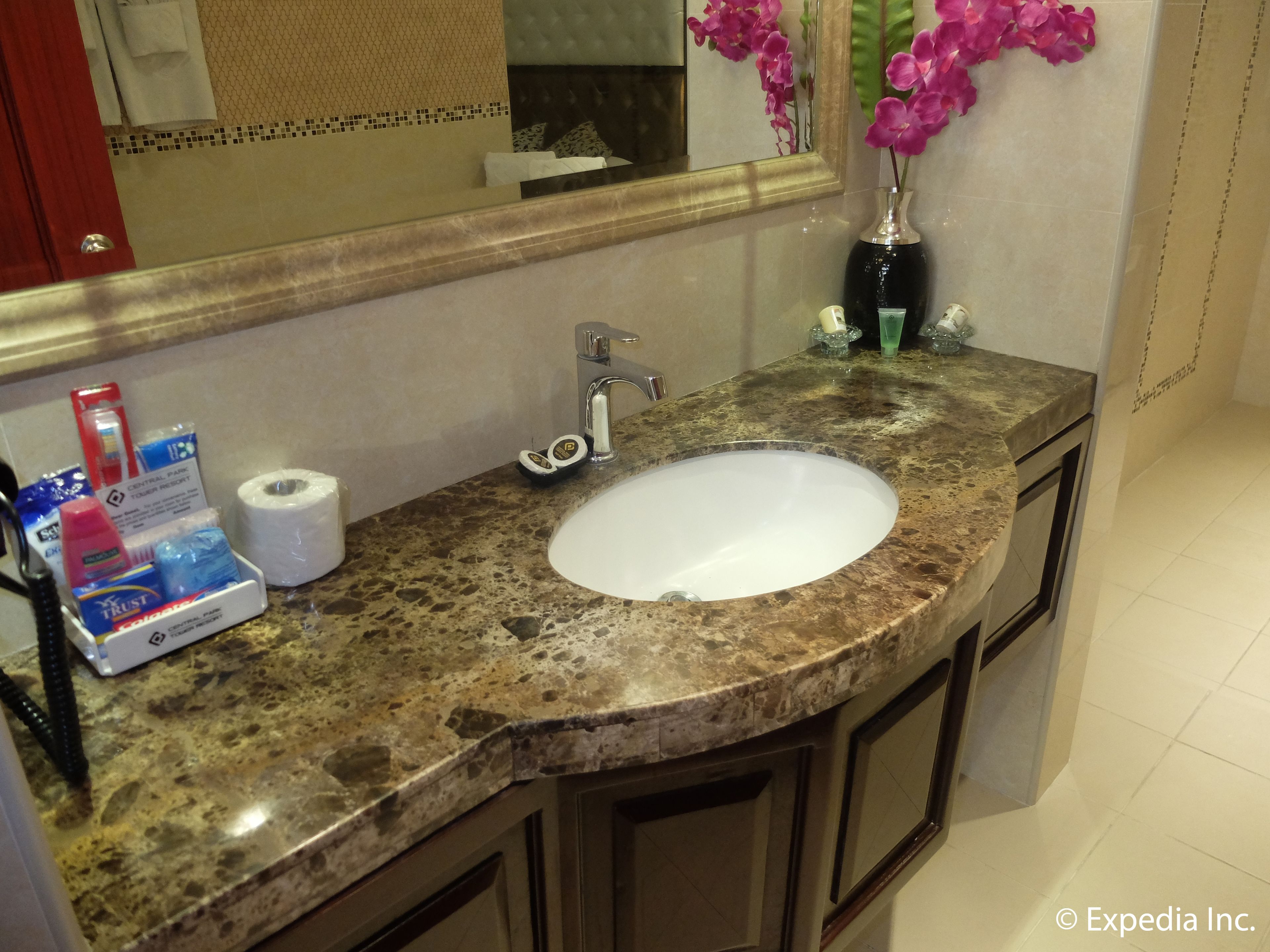 A bathroom with a marble sink, a mirror, a vase with pink flowers, toiletries, and a towel rack.