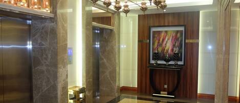 A modern lobby with marble flooring, a chandelier, elevators, and a wall-mounted television.