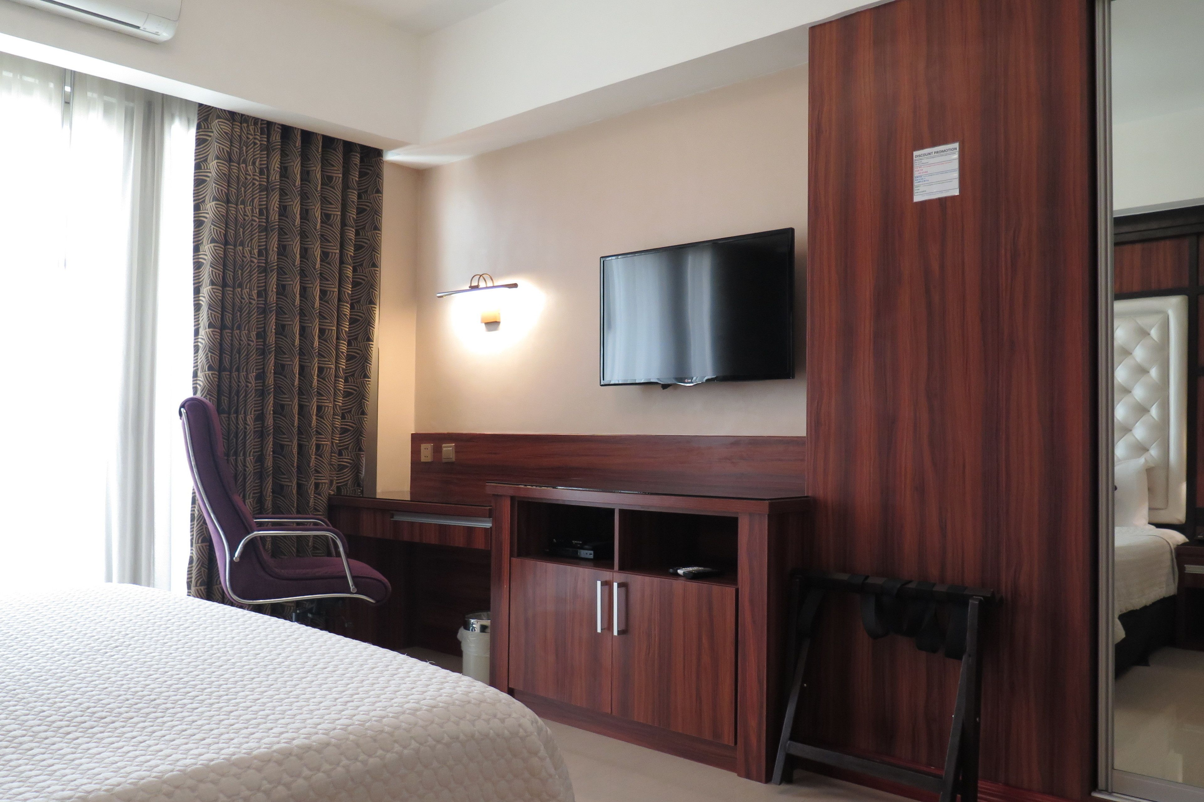 A hotel room with a bed, a desk with a chair, a television mounted on the wall, and a wardrobe.