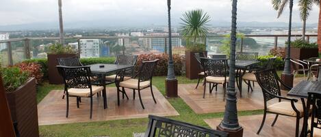 An outdoor patio with tables and chairs, surrounded by greenery and a city view.