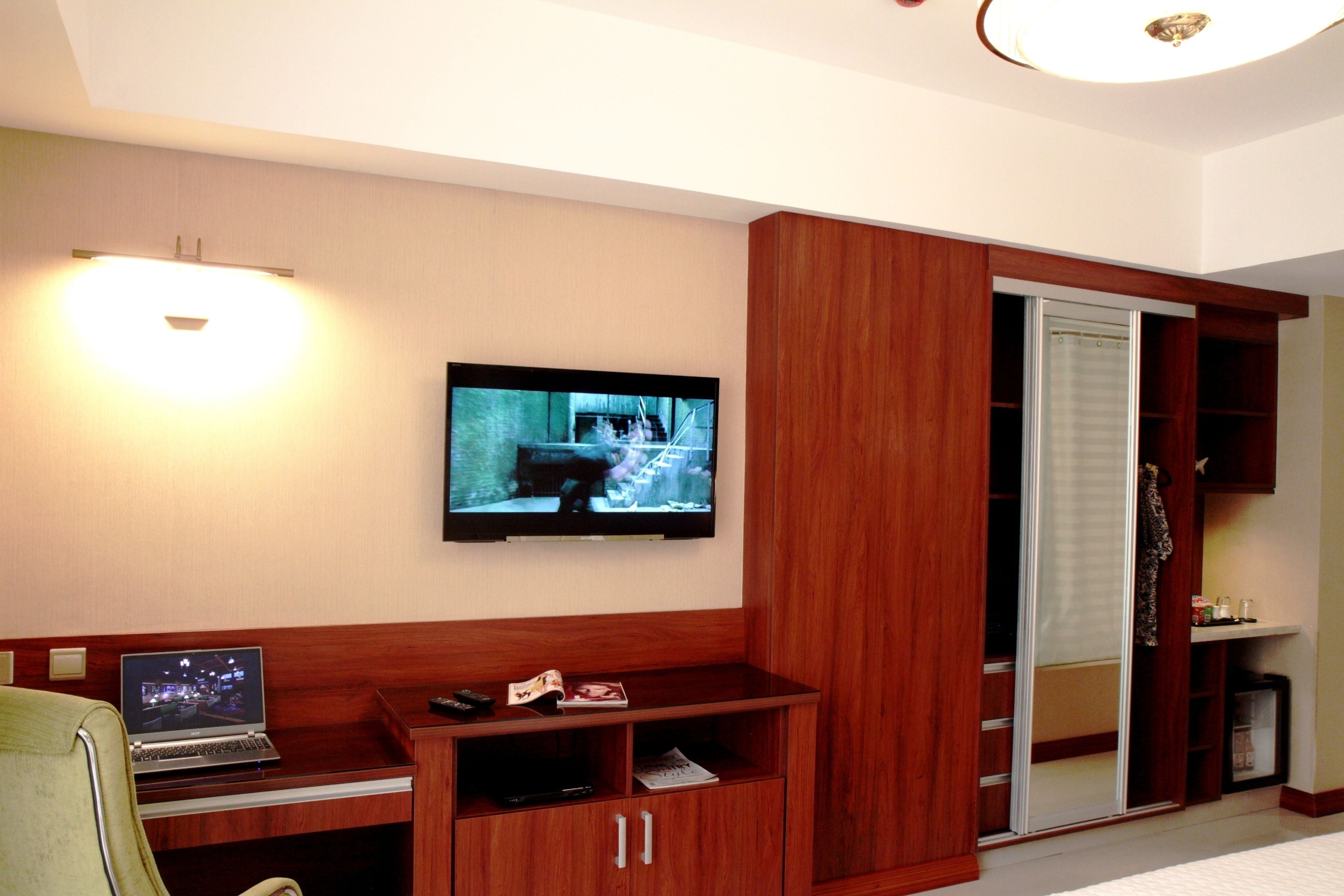 Una habitación con una televisión de pantalla plana montada en la pared, un ordenador portátil sobre un escritorio de madera, un armario de madera y un armario con puerta de vidrio corredera.