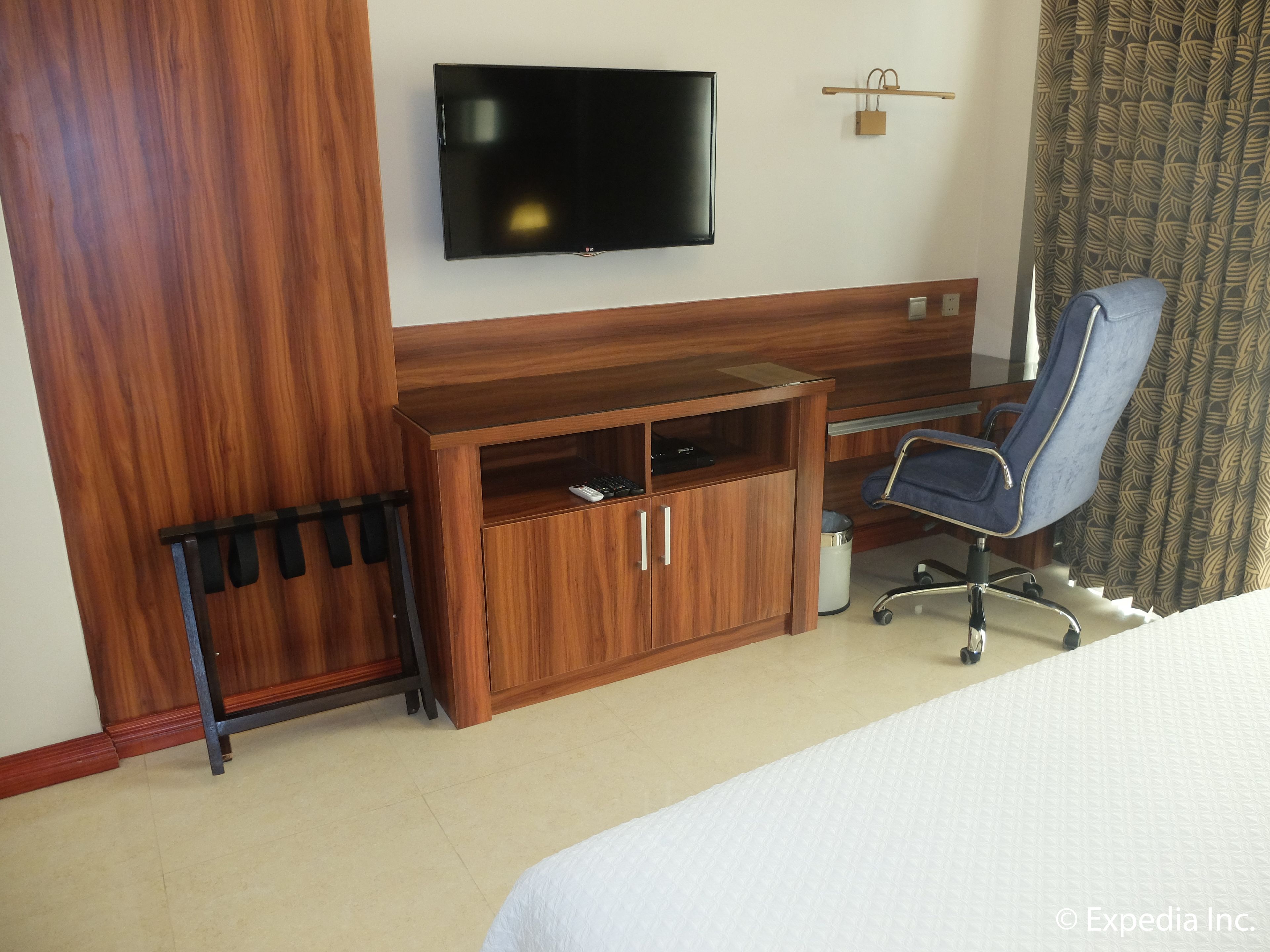 A hotel room with a wooden desk, a television mounted on the wall, a chair, and a bed.