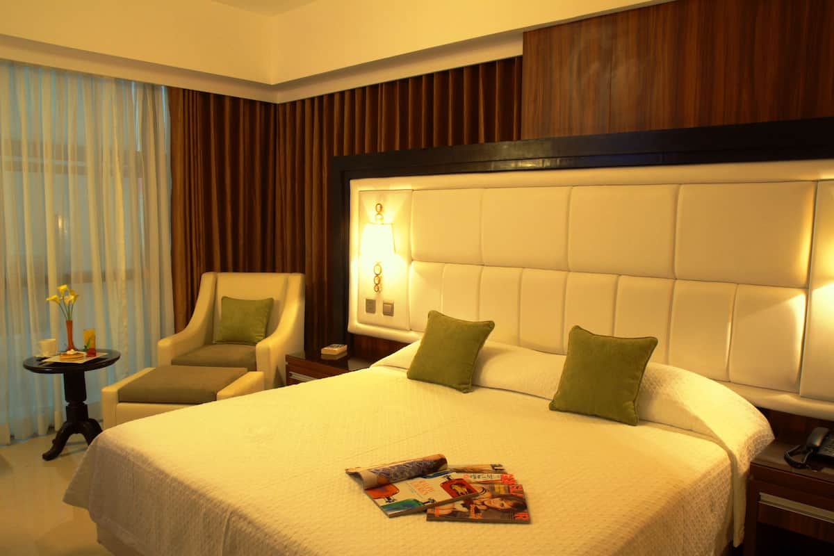 Ein Hotelzimmer mit einem großen Bett, zwei Kissen, einem Nachttisch mit Lampe, einem Sessel, einem kleinen Tisch mit Vase und Zeitschrift sowie einem Fenster mit Vorhängen.